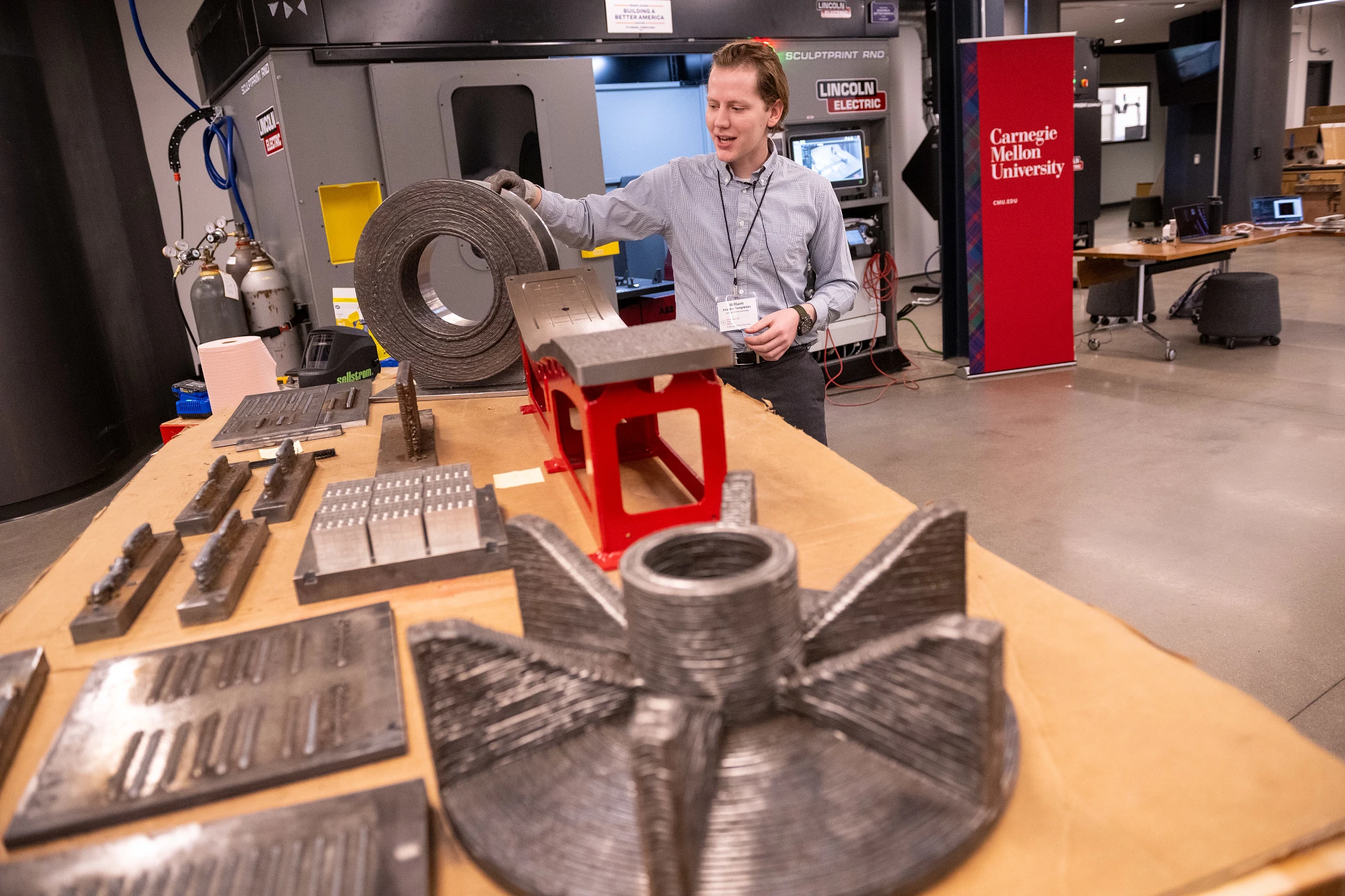 A Carnegie Mellon researcher demonstrates metal 3D-printed parts on a workshop table, with a large industrial printer behind him; the scene feels hands-on and innovative.