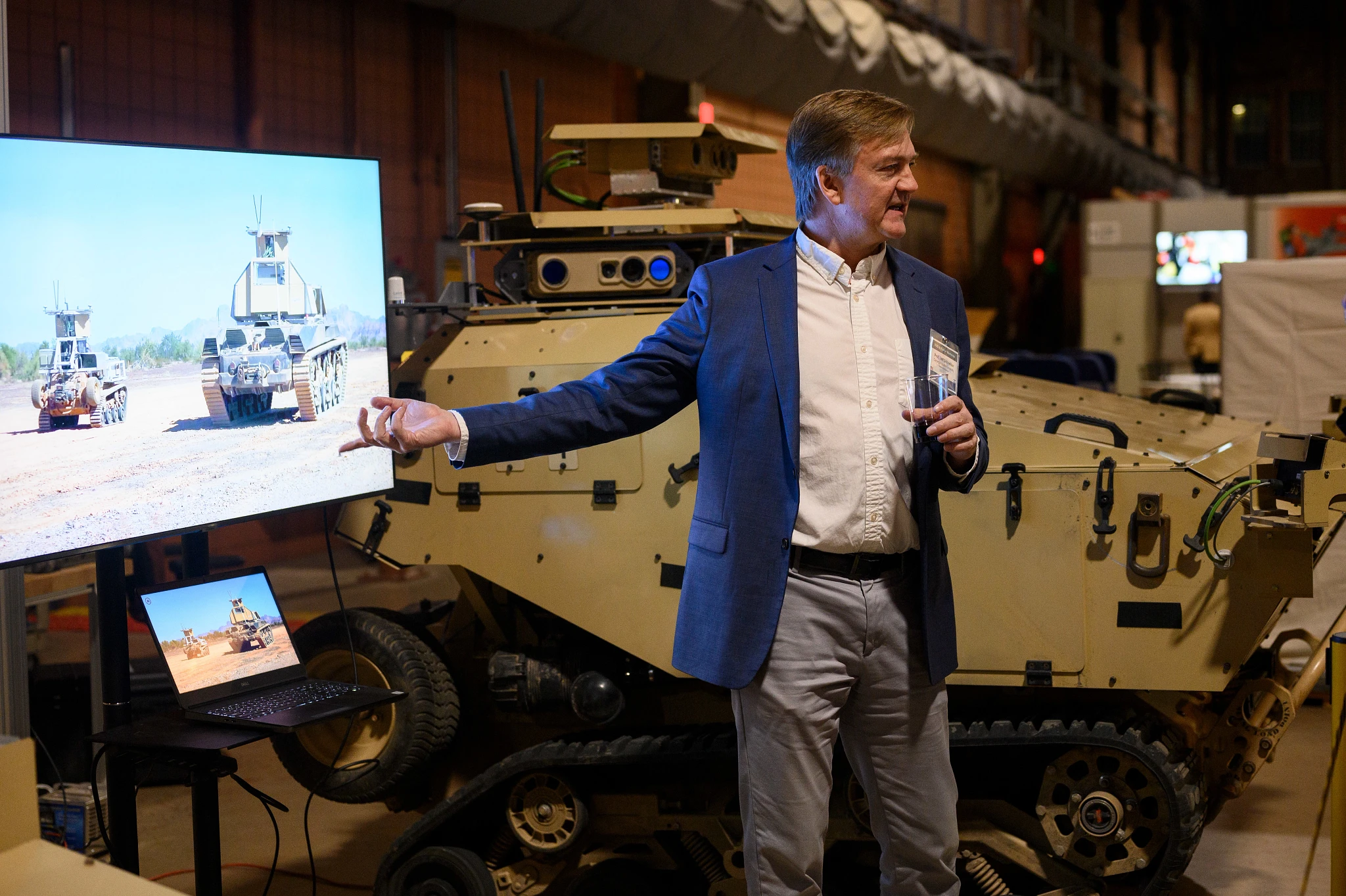A man in a blue blazer gestures toward a screen showing military vehicles, presenting beside a tan armored vehicle at an indoor exhibition.