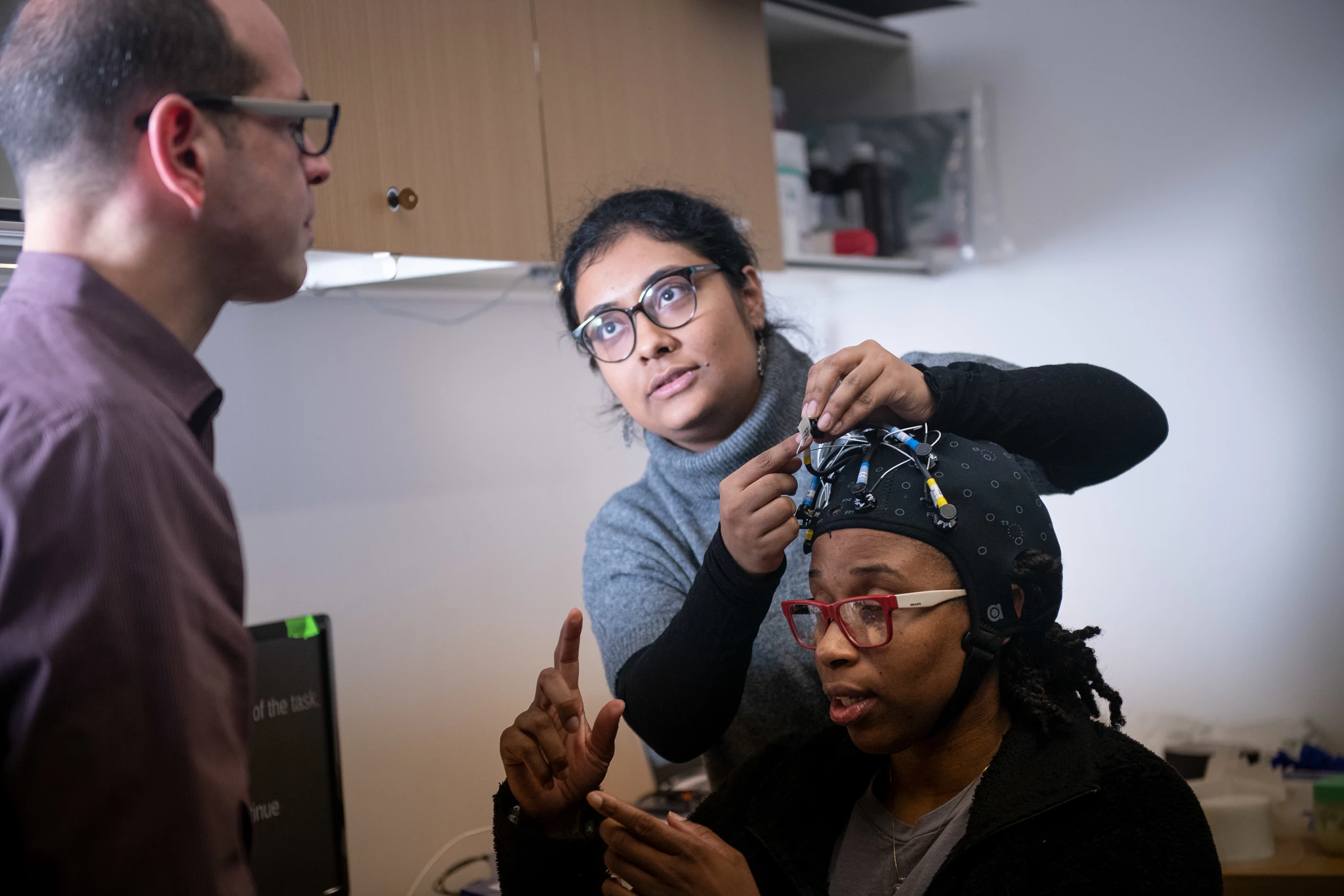 A researcher adjusts an EEG cap on a seated participant while a colleague watches in a lab, creating a focused, collaborative neuroscience research scene.